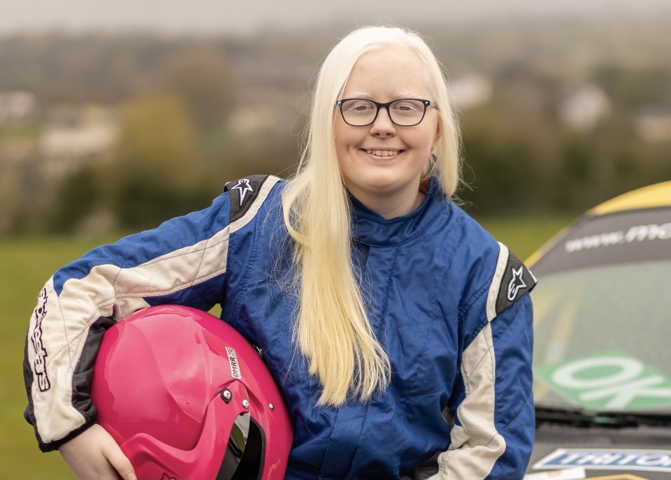 a girl with long white hair stands in front of a yellow rally car holding a pink race helmet