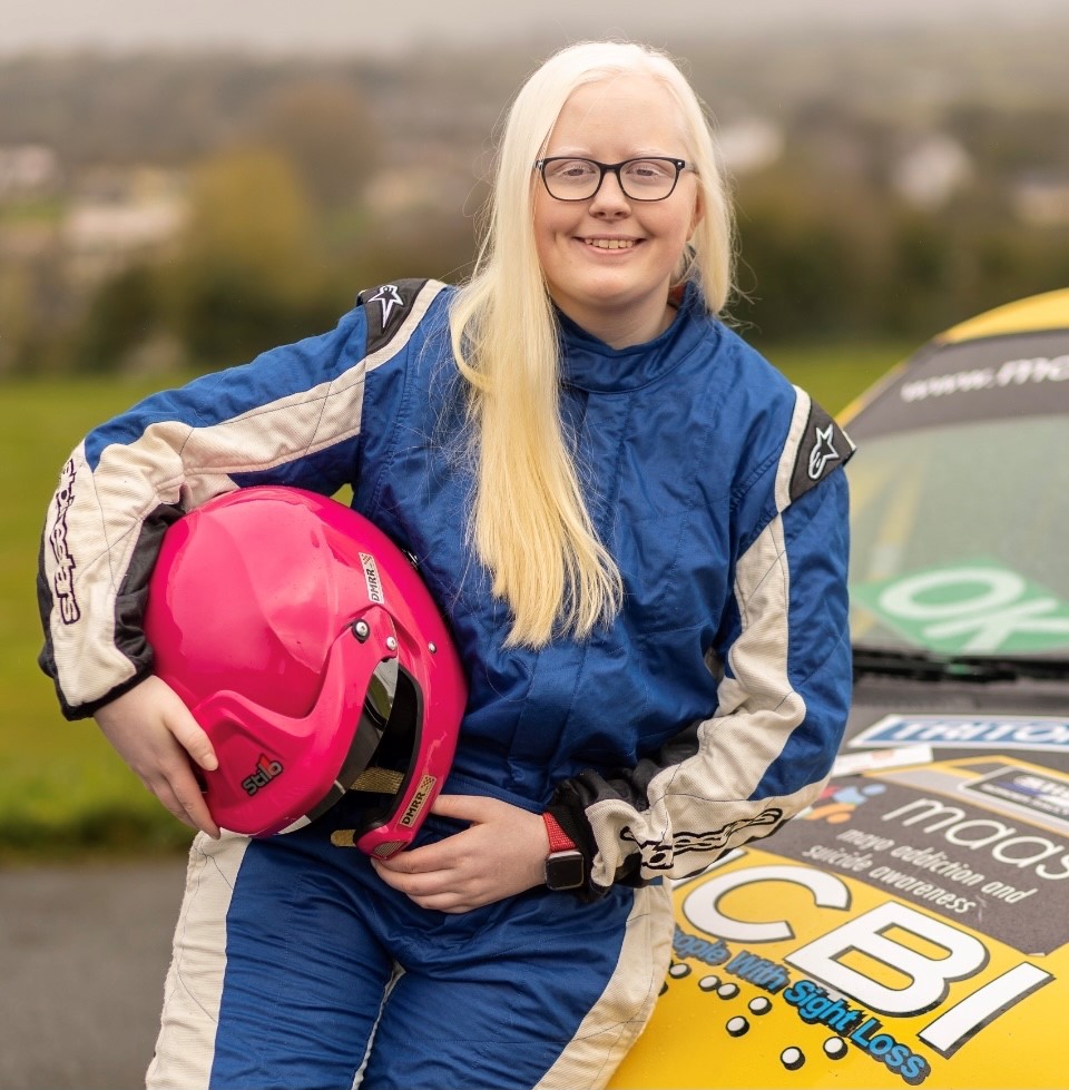 a girl with long white hair sits on the front of a yellow and black rally car holding a pink rally helmet
