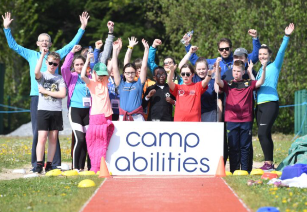 a group of children and adults stand behind a camp abilities sign with their arms raised cheering