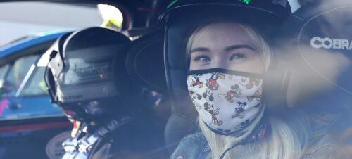 a girl with blonde hair sits in a rally car wearing a face mask and rally helmet