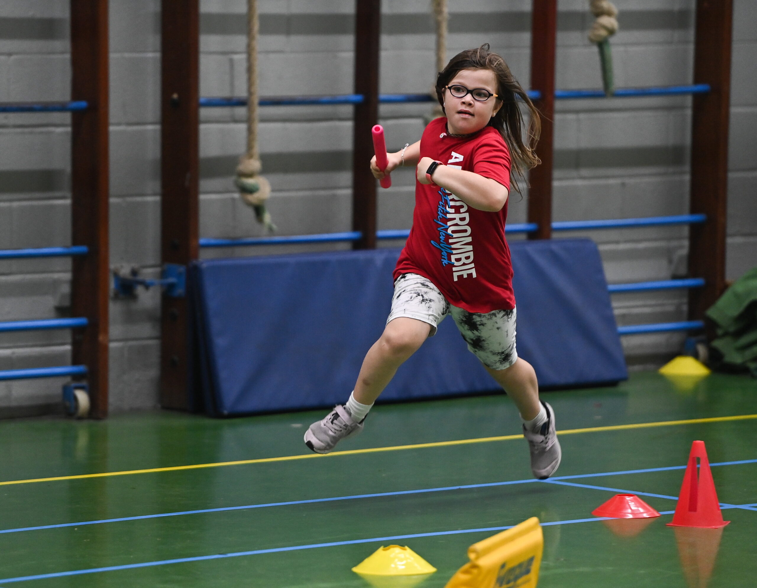A young girl wearing glasses takes part in a track race and she is holding a red baton.