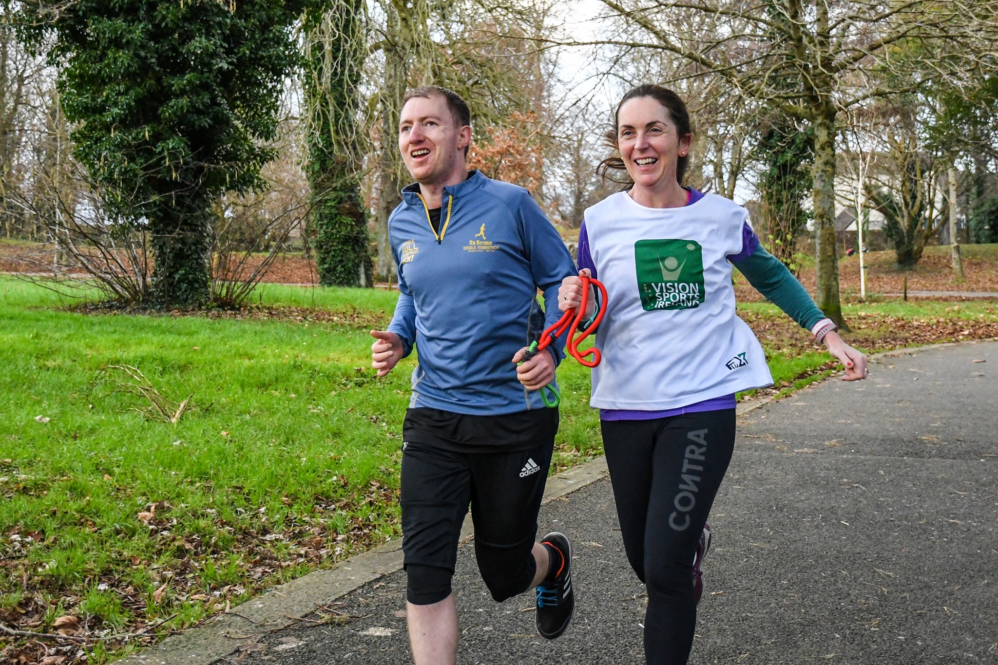 A woman and a man run together through a park area. The man has a red rope tied around his arm which the woman, who is wearing a Vision Sports Ireland t-shirt, is holding.