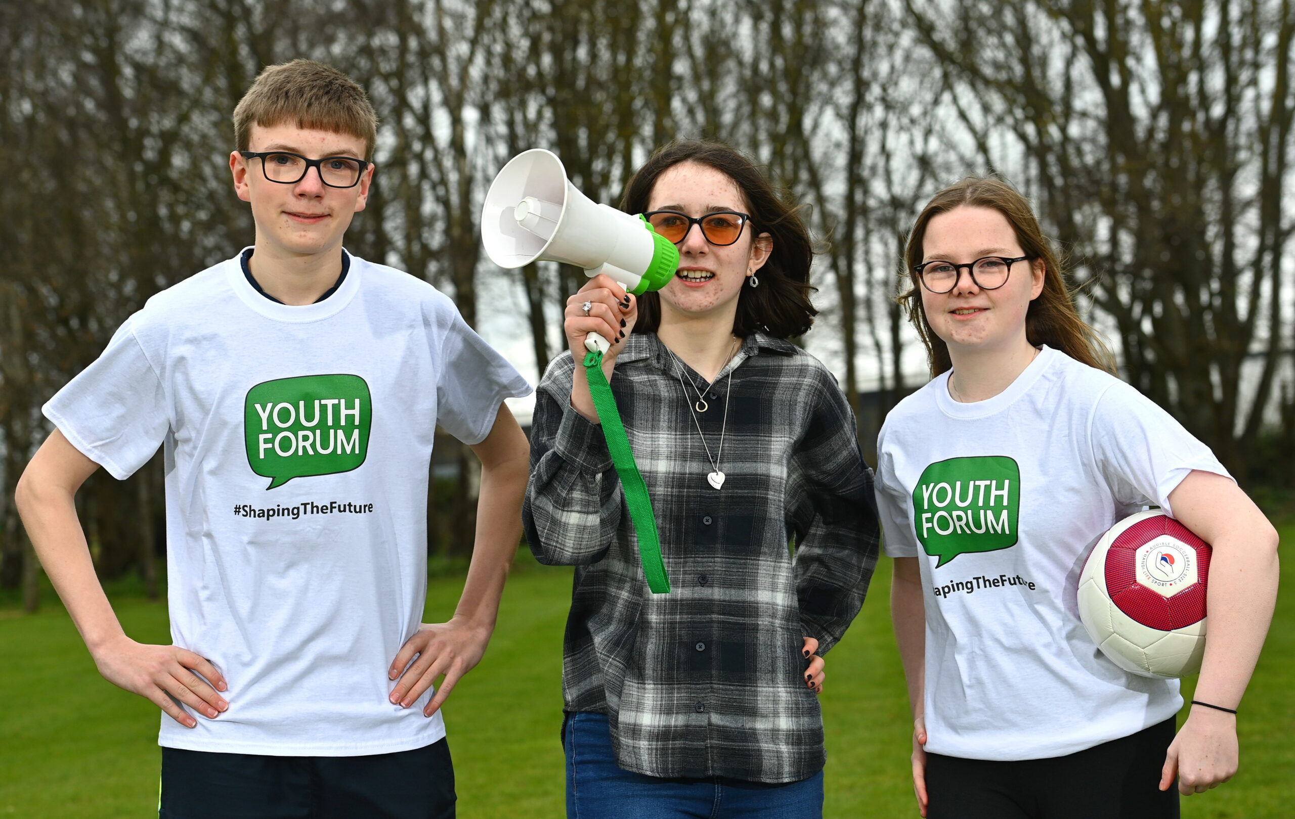 One boy and two girls stand together. The girls are holding a megaphone and a football while the boy stands with his hands on his hips. The boy and one girl are wearing white t-shirts with the Youth Forum logo on the front.