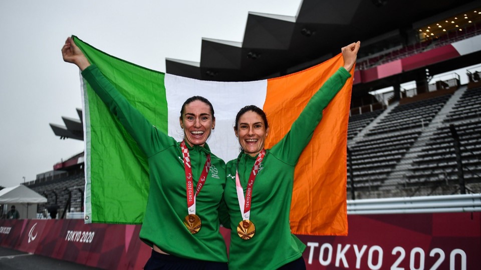 Eve and Katie George standing on the podium grinning iwth an Irish flag wearing a gold medal