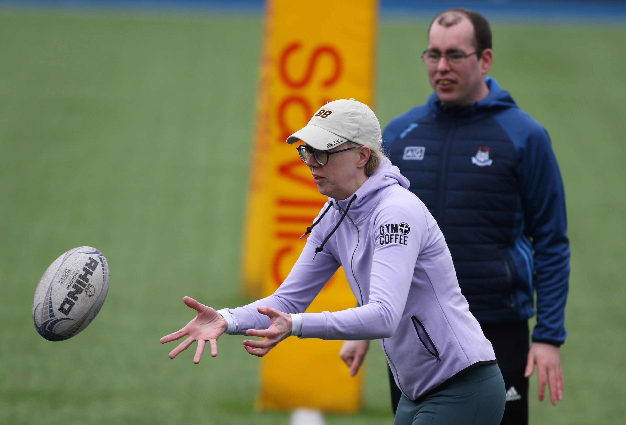 A man stands behind a woman who is wearing a cap and glasses who is about to catch a rugby ball which is coming toward her in a mid-air