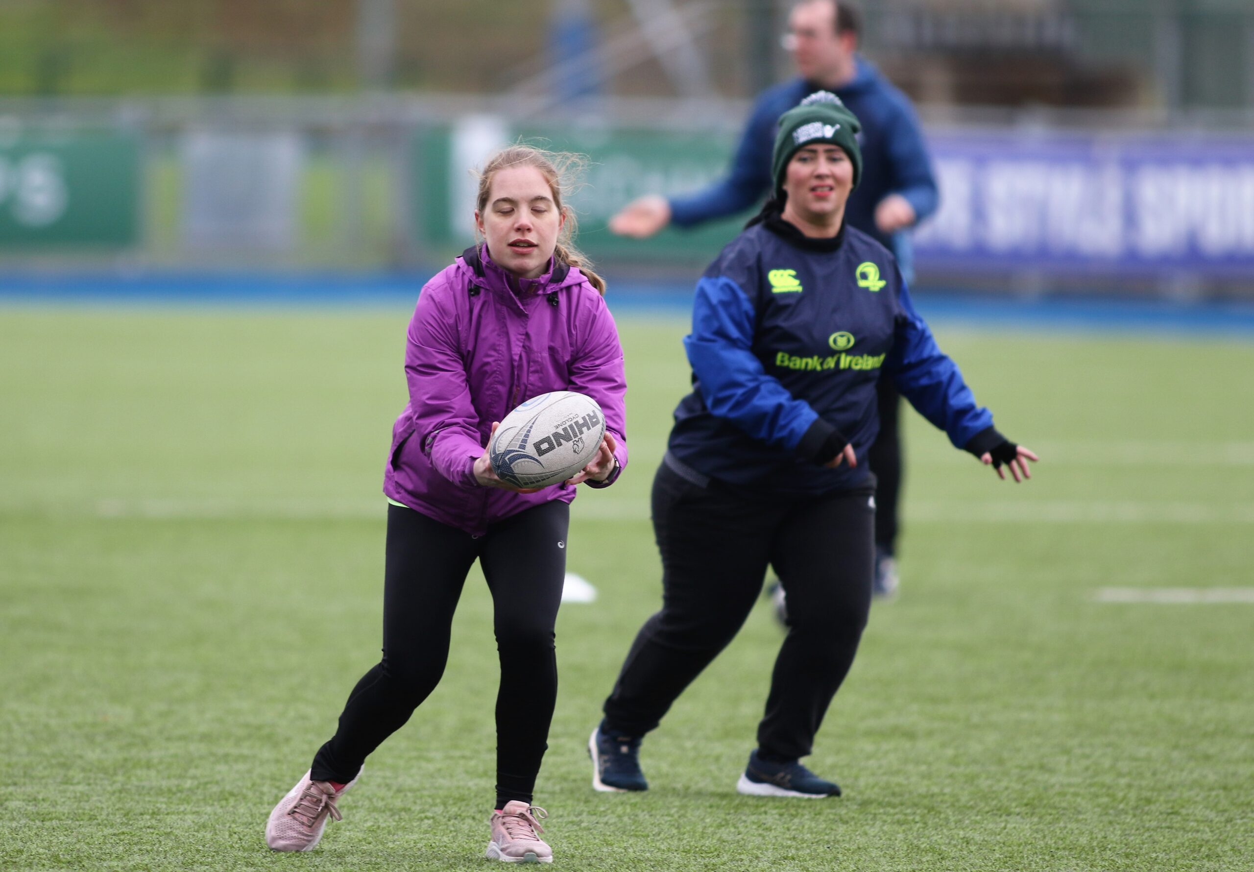 A young woman who is wearing a purple top catches a rugby ball, while another woman who is wearing a Leinster training outfit runs in behind her ready to receive a pass.