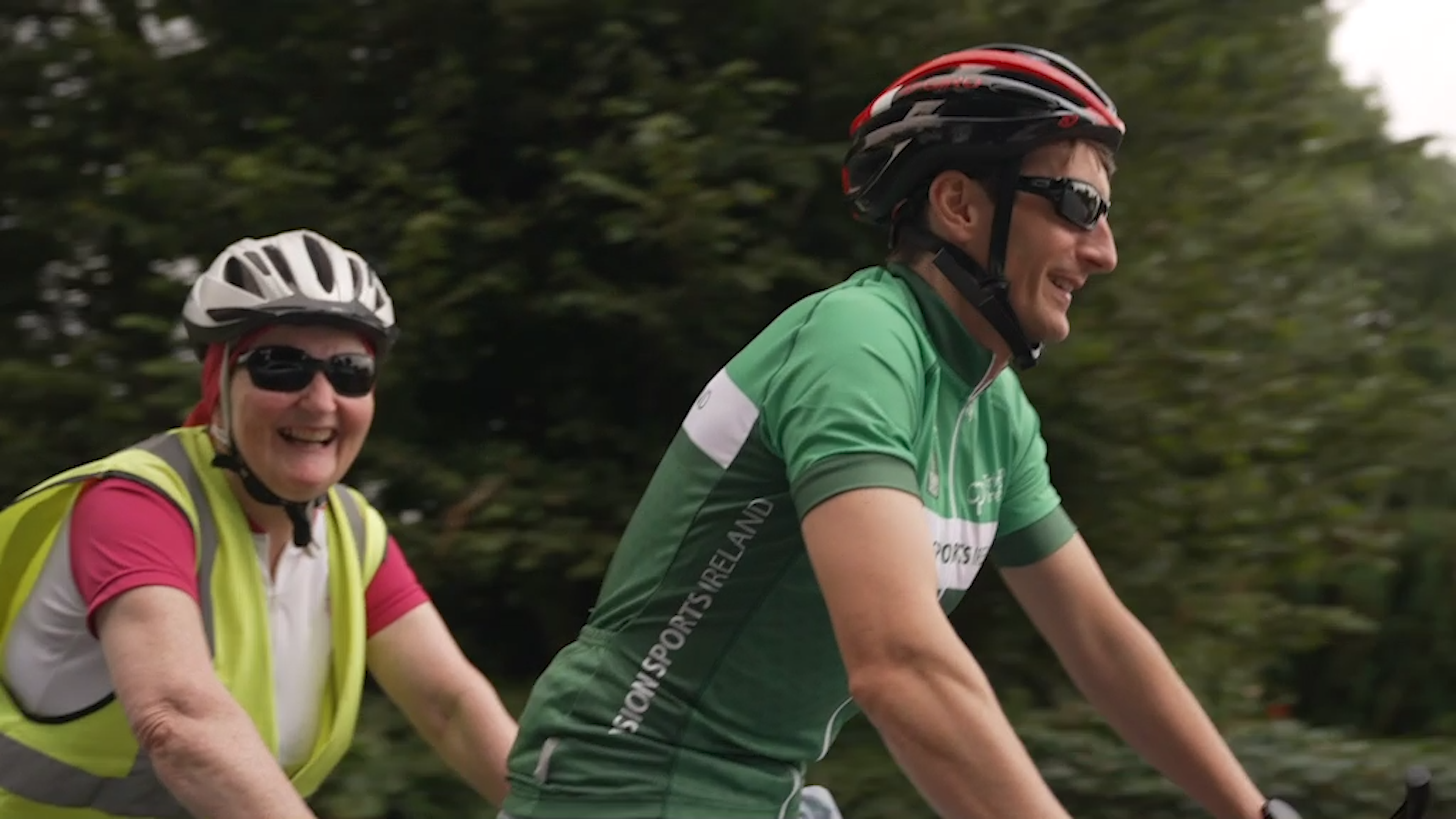 A woman and a woman, both of whom are wearing high visibility vests and sunglasses, smile as they are photographed while cycling