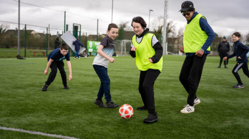 Young players playing football, two are wearing high vis vests and one is kicking a football