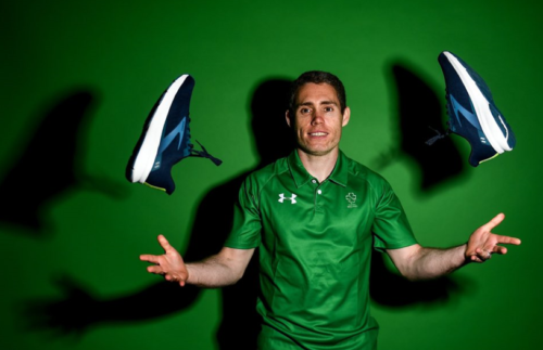 Image of a man standing in front of a green background wearing a green Paralympics top throwing a pair of trainers in the air