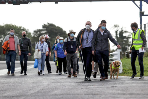 a large group of participants and volunteers walking towards the camera some are using canes and are receiving sighted guide There is a guide dog in the corner of the screen