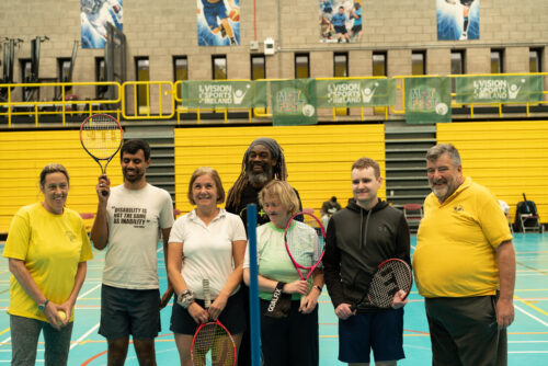 Photo of a group of five athletes, two male and three female with three tennis coaches, holidng tennis racquets