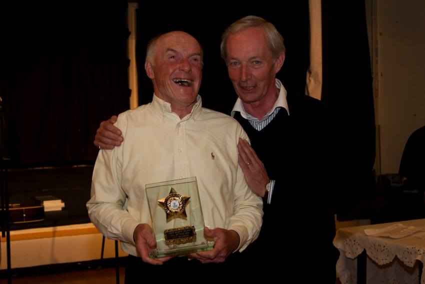 two men one with their arm around the other pose for a photograph. One is holding a trophy with a gold star on it