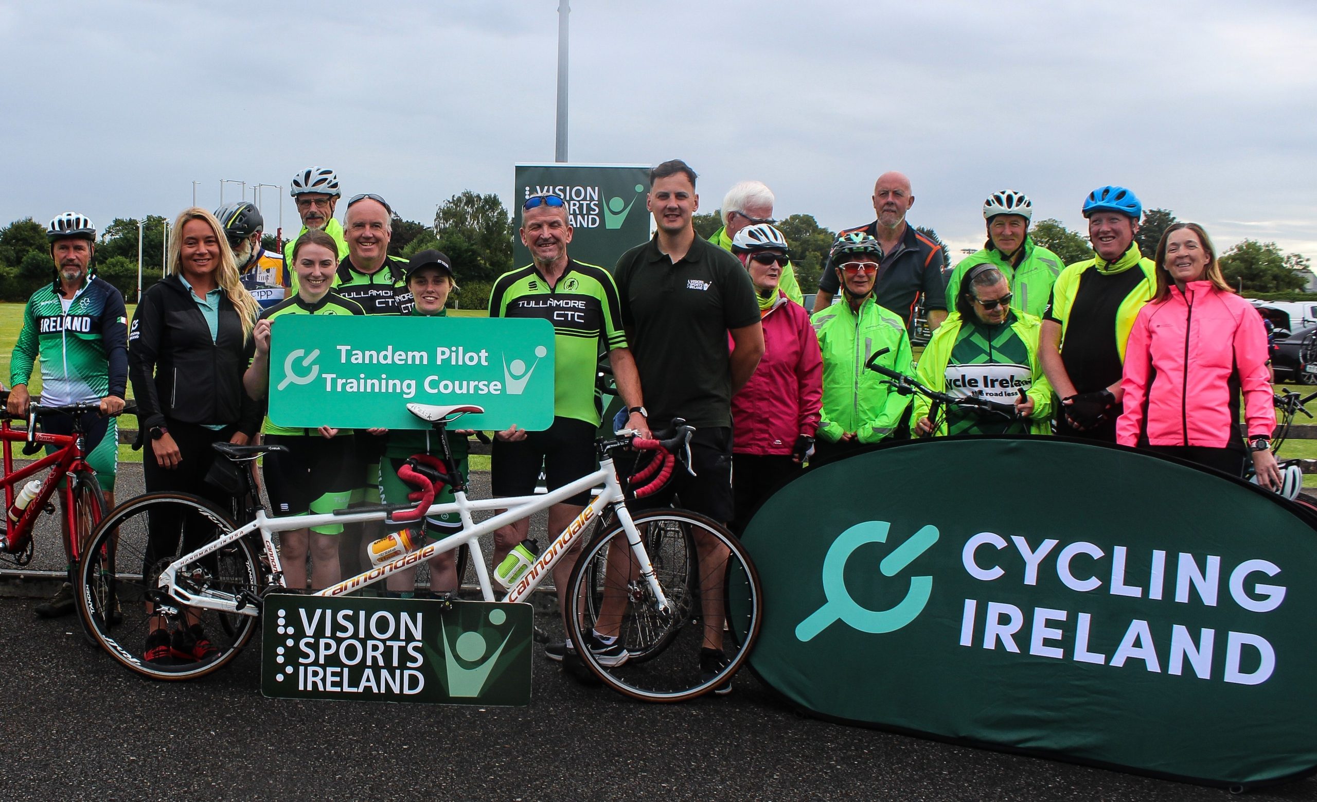 Group photo of tandem pilots and stokers holding up signs with VSI, tandem pilot training course and cycling Ireland logos . There are also some tandem bikes standing in front of the people