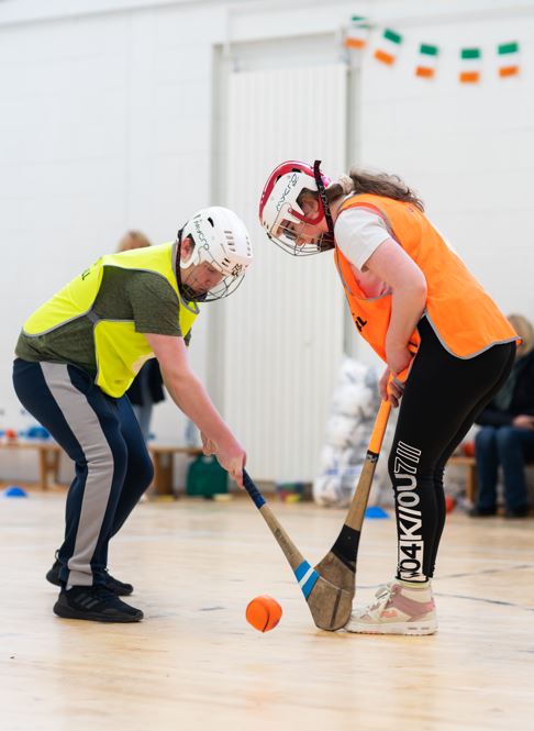 2 teens wearing bibs and hurling helmets, hold hurls and contest a large orange ball on the floor