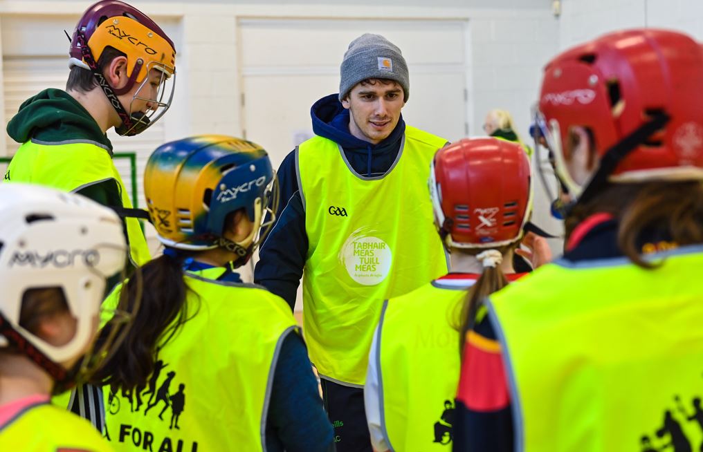 a group of teens wearing yellow bib standing around a male limerick hurling player who is wearing a woolen hat