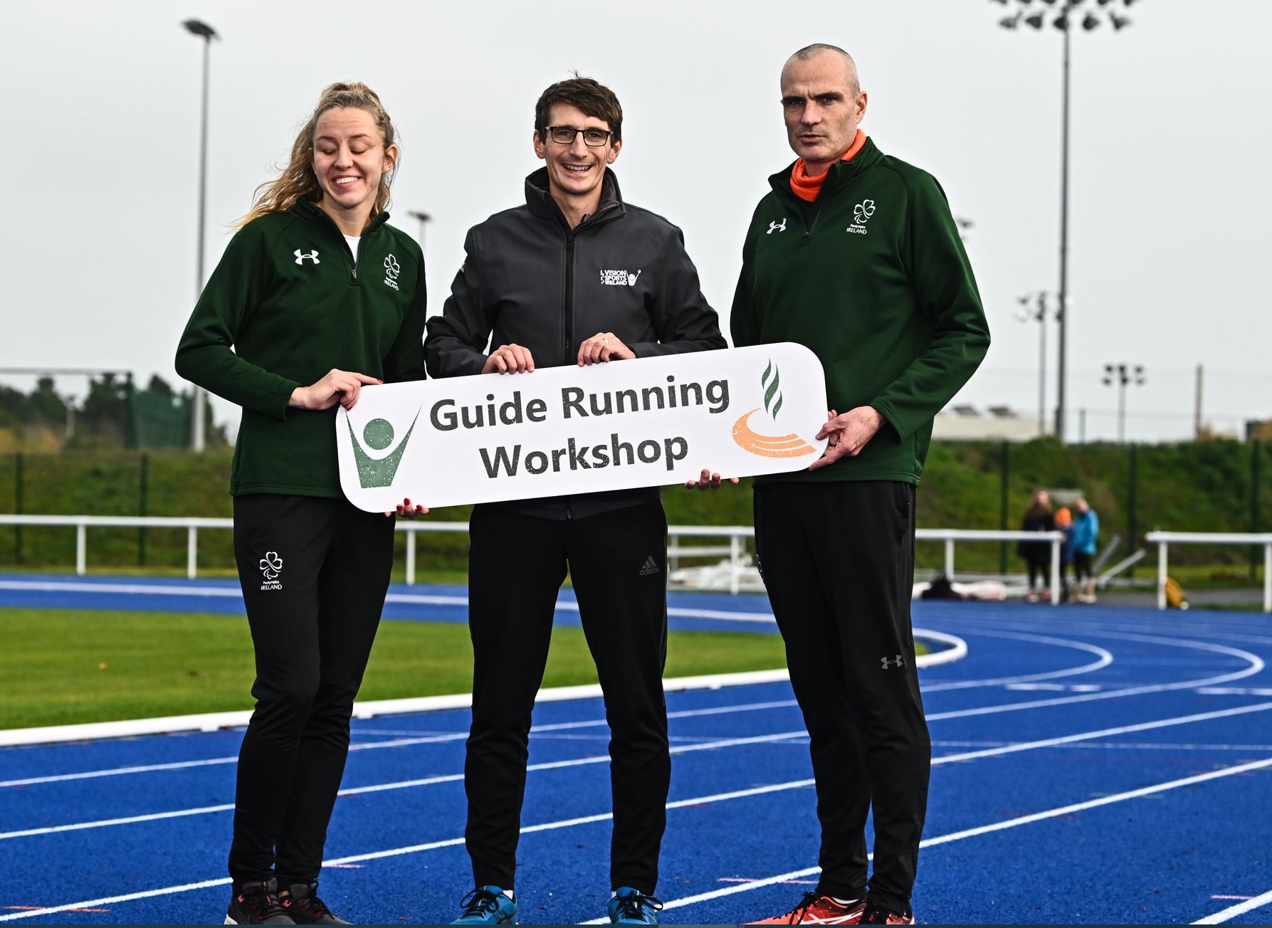 3 people stand on a blue running track holding a white sign saying guide running workshop