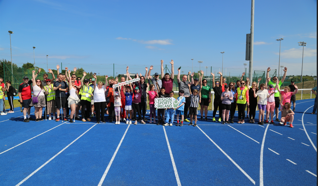 Athletics Group – Athletes and guides cheer as a group holding a Vision Sports Ireland and Get Active Everywhere signs while standing on the blue UCD athletics track.