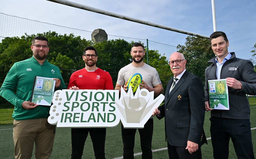 A group of 5 males including Robbie Henshaw and Ian McKinley stand for a photograph holding VI Rugby coaching guides, rugby balls and a Vision Sports Ireland logo sign