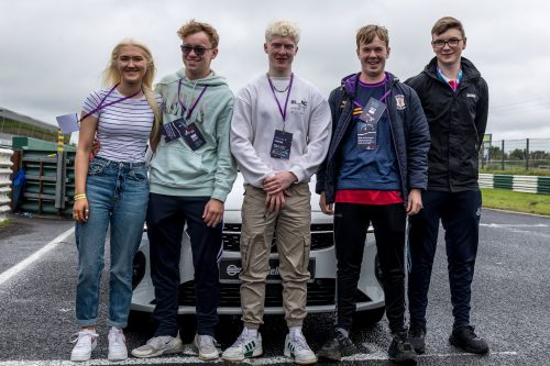 A group of participants stand in front of a car, smiling at the camera after driving around the track.