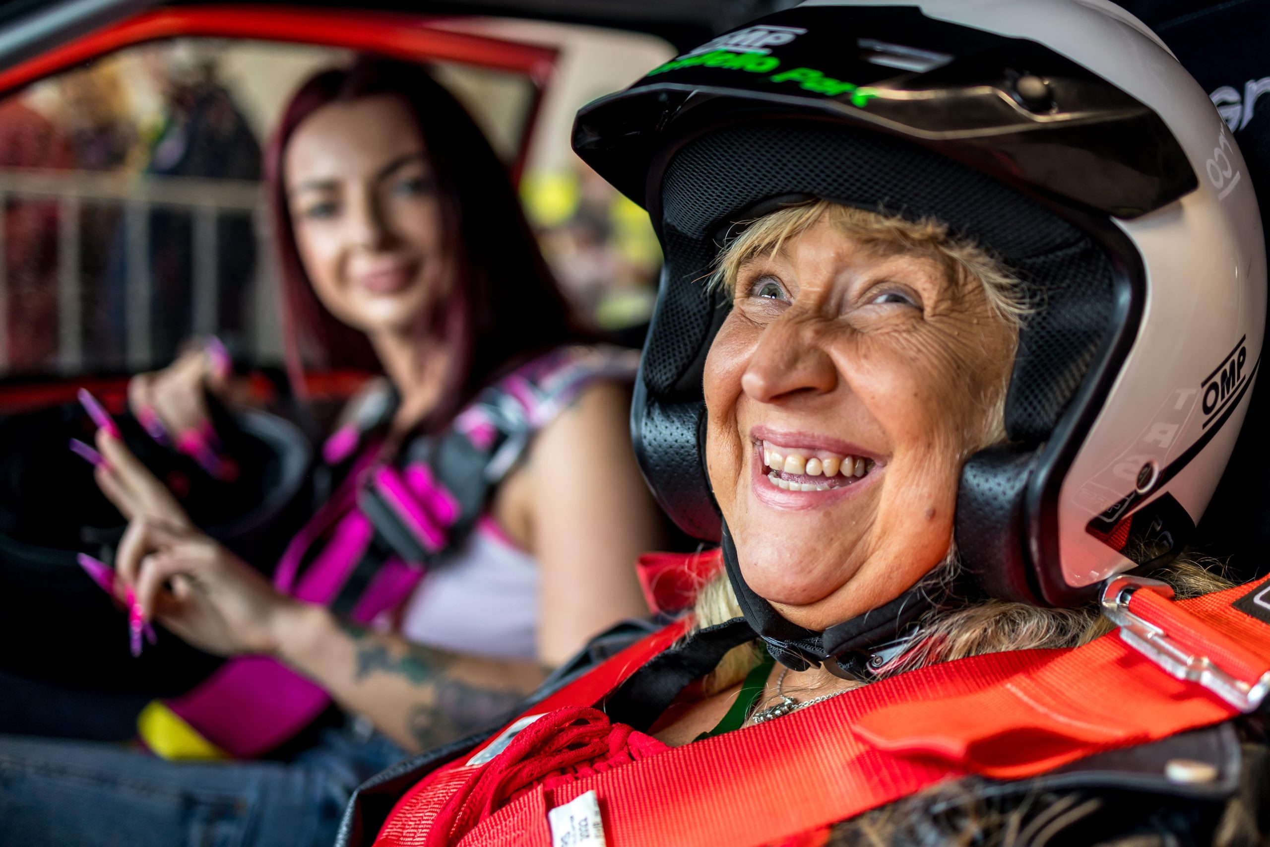 A participant wearing a helmet and sitting in the passenger seat of a rally car, smiling at the camera at #ZeroLimits23. The rally driver is visible in the background.