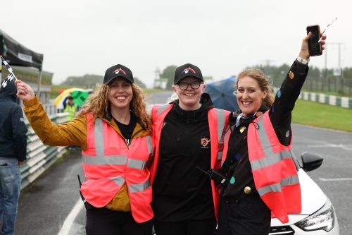 Three staff members in pink high vis jackets smile and wave at the camera standing in front of a car on track