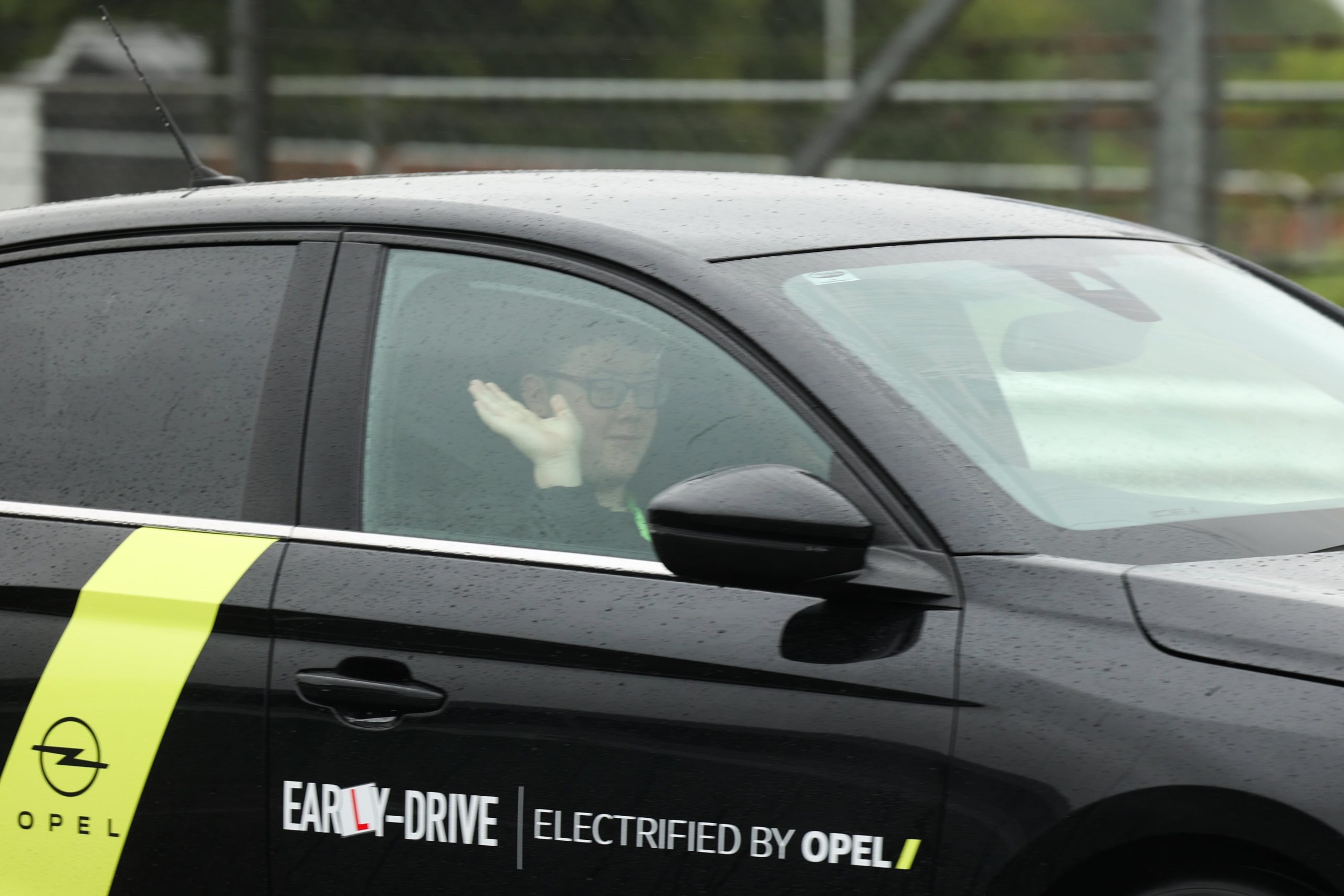A paticipant at #ZeroLimits23 drives a black car on track while waving to the camera.