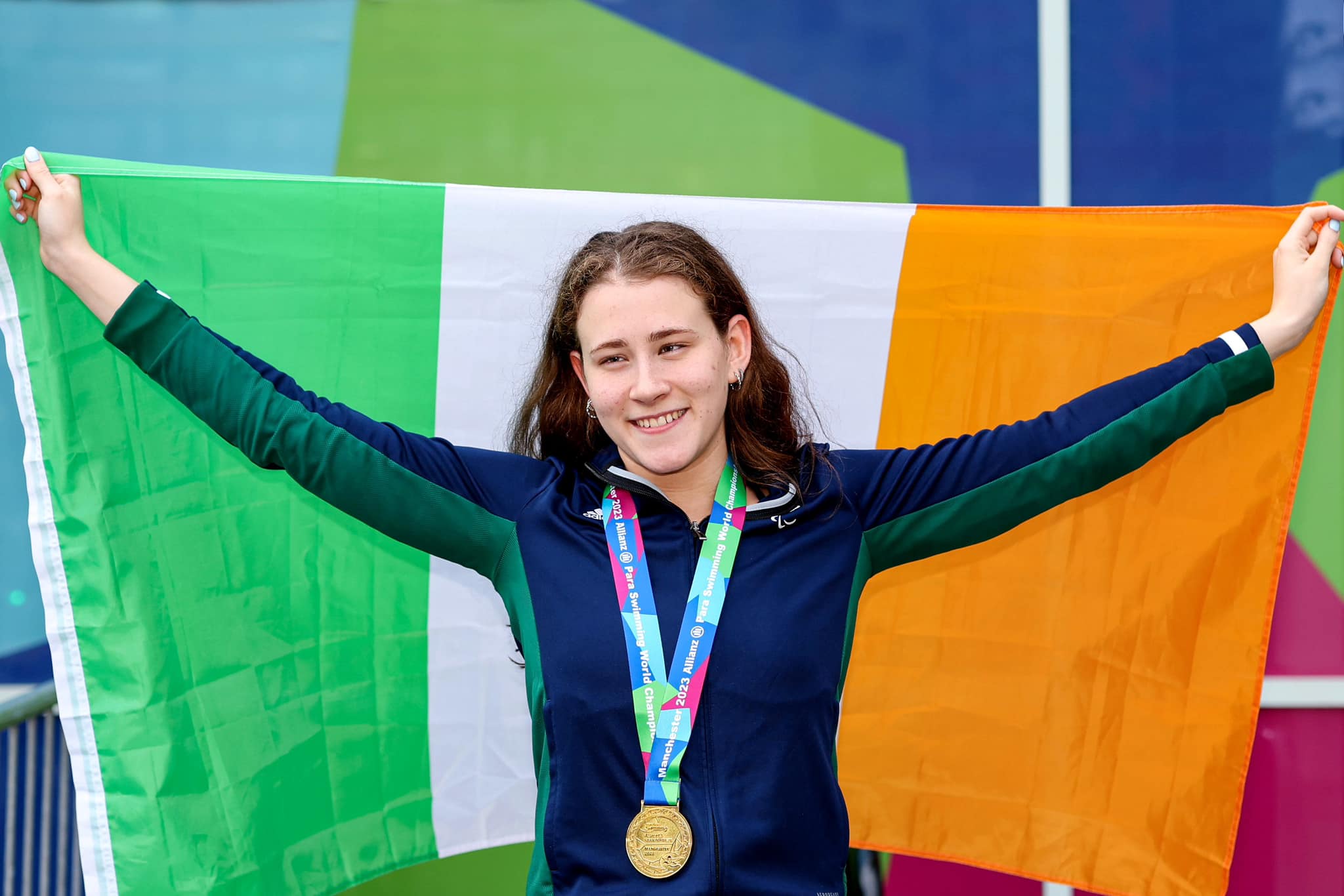 Roisin Ni Riain stands holding an Ireland flag behind her and a gold medal around her neck