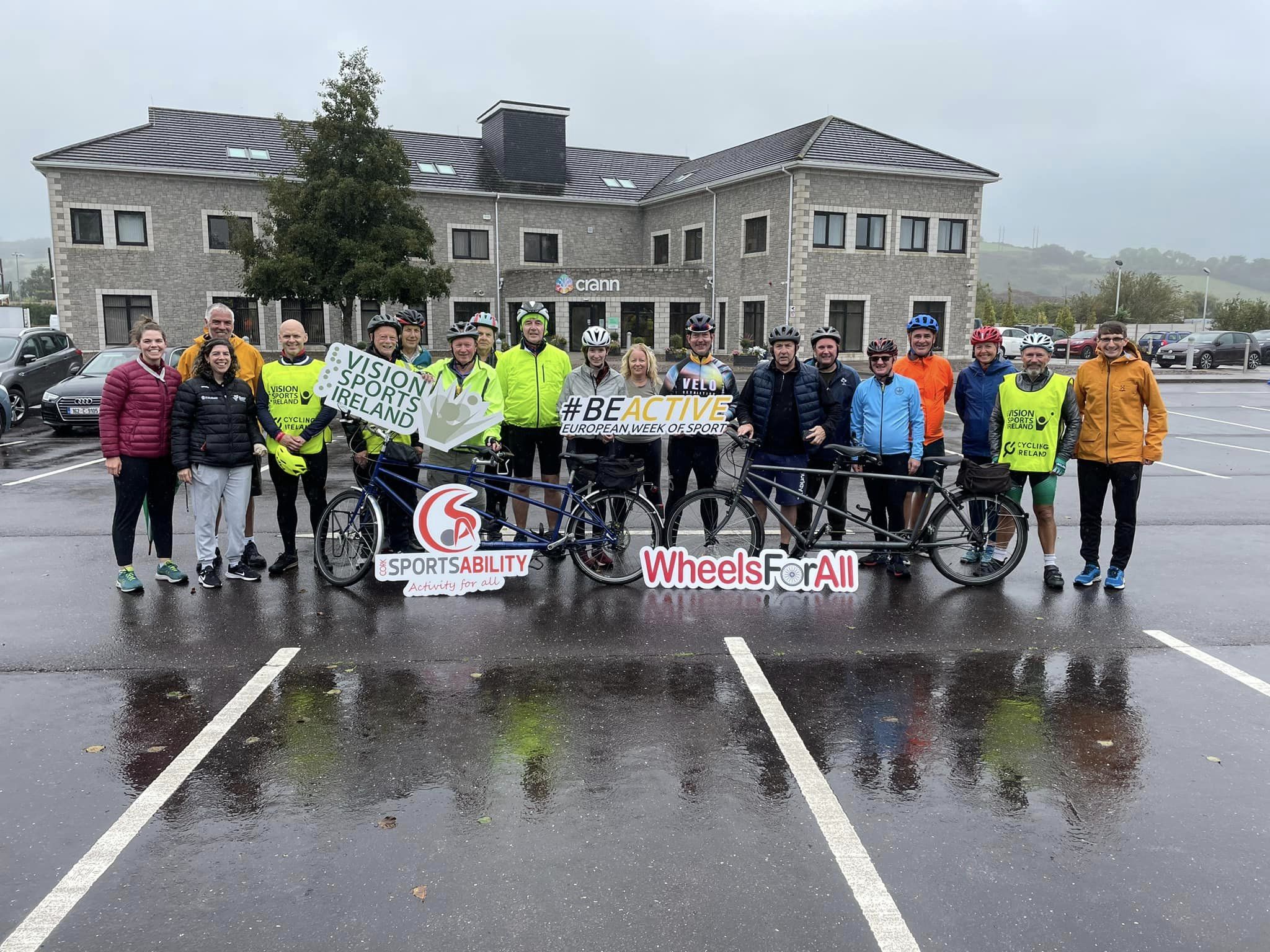 A group of participants and guides at a tandem pilot course. The group are looking at the camera and holding signs for Vision Sports Ireland, Cork SportsAbility, Wheels for All and #BeActive European Week of Sport.