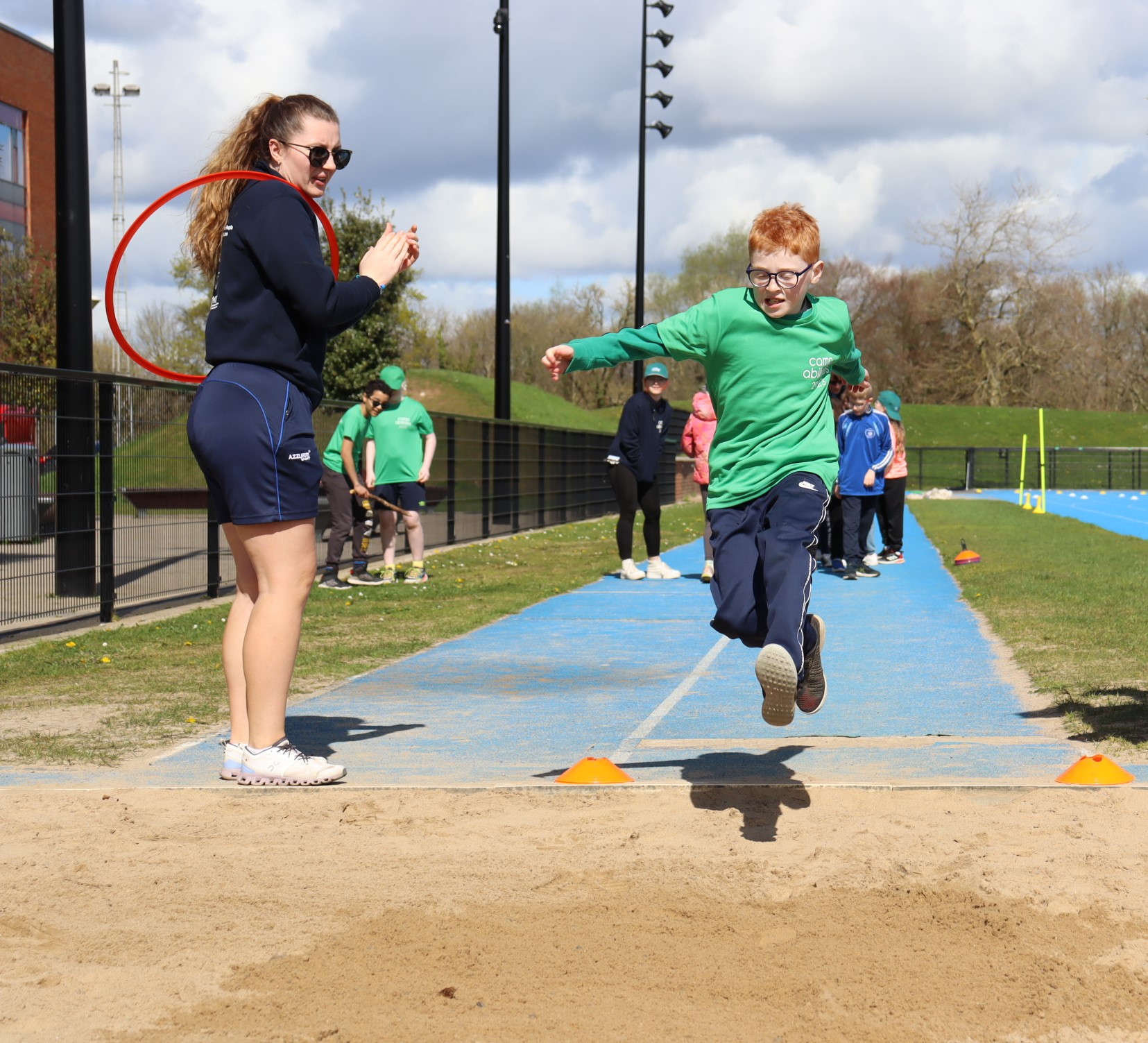 Female Coach claps as young red haired boy completes long jump on a blue running track into yellow sand.