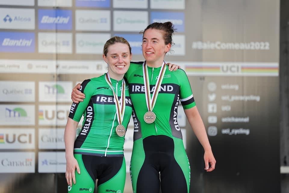 Josie, a female with her hair tied up and her tandem pilot stand on a winners podium with medals around their necks
