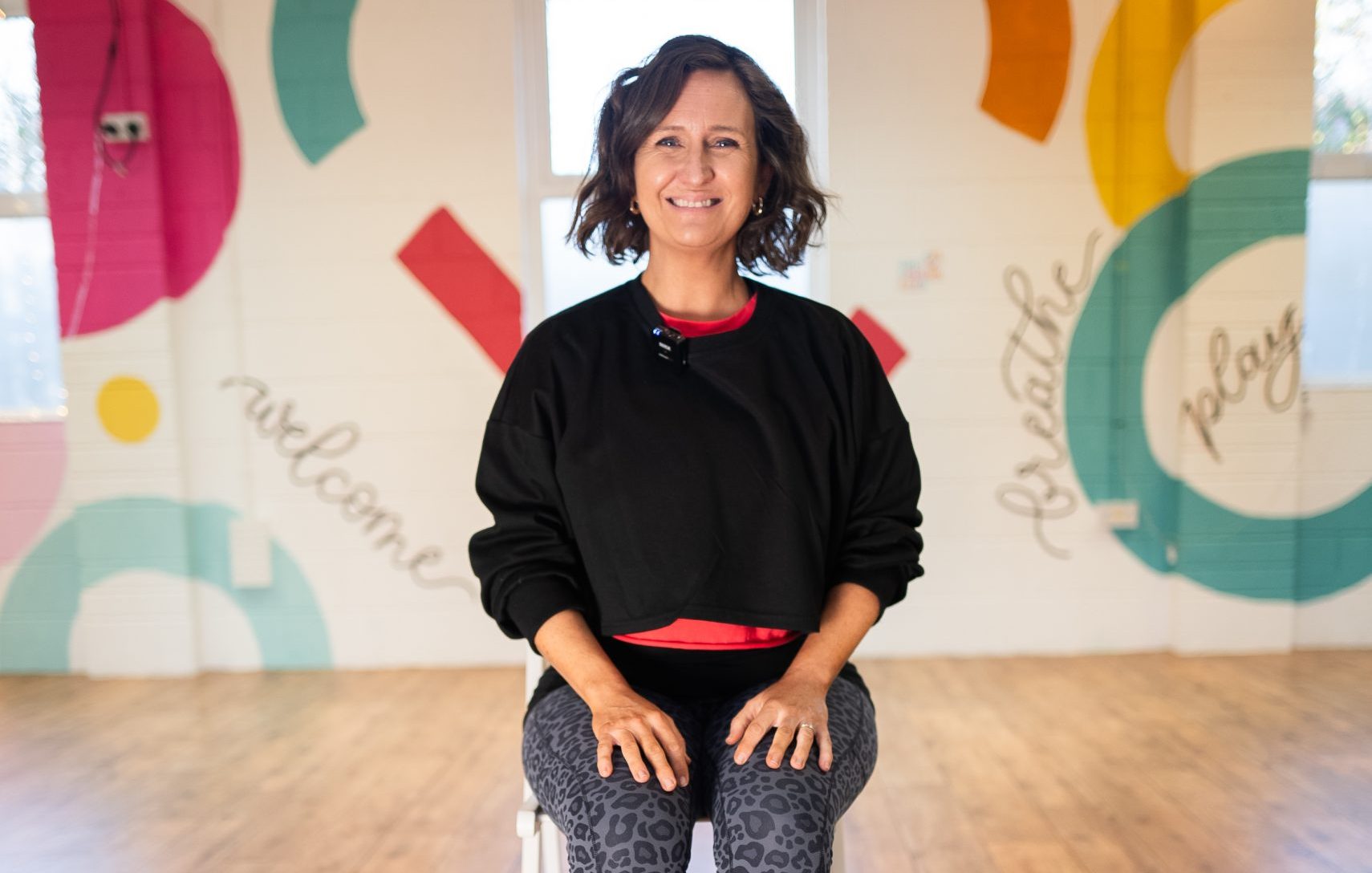 Yoga instructor Siobhan sits on a chair with a mat behind her in a colourful studio